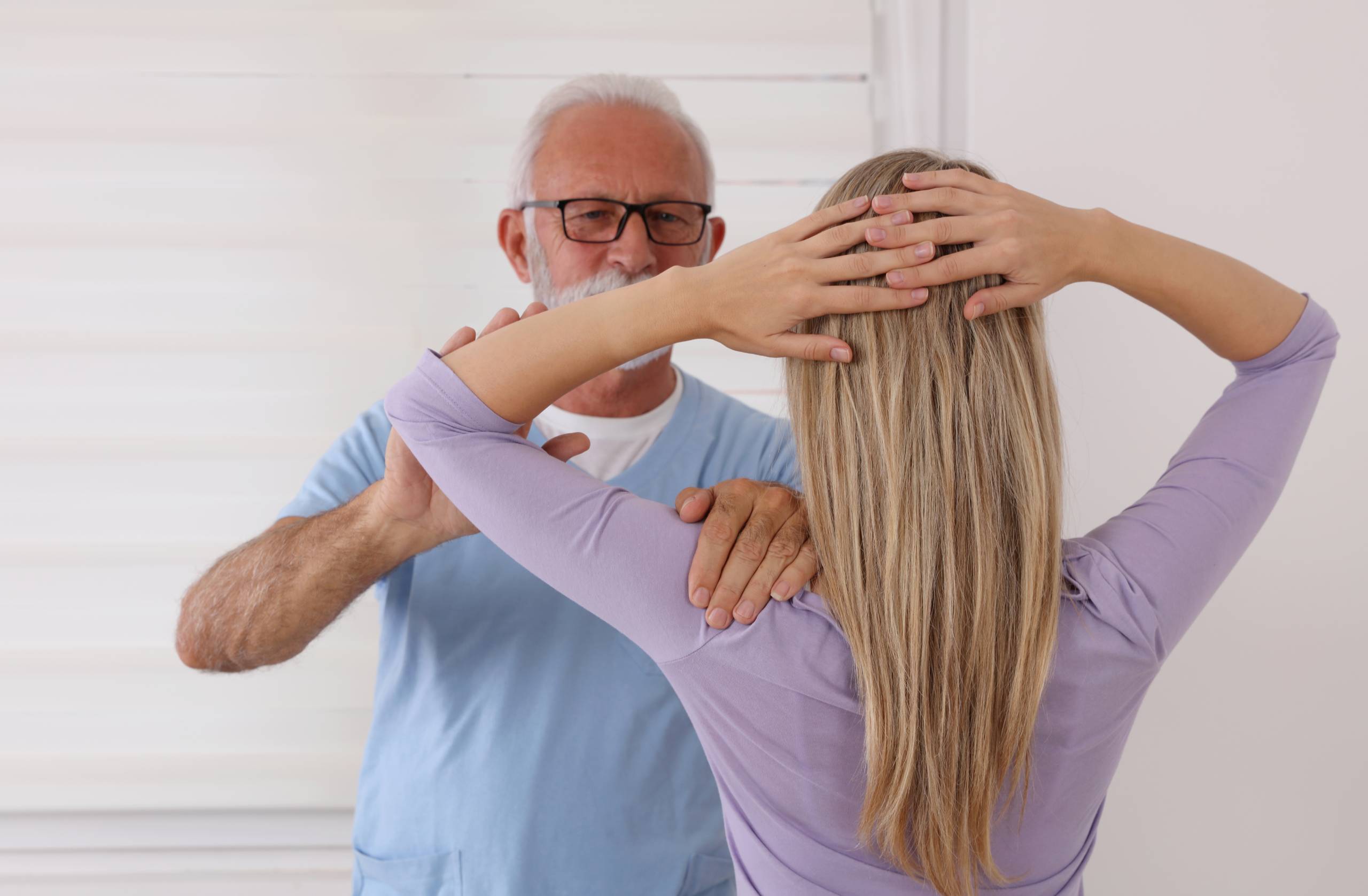 A physical therapist in blue scrubs assesses a blonde female patient during a rehabilitation or corrective care exercise.