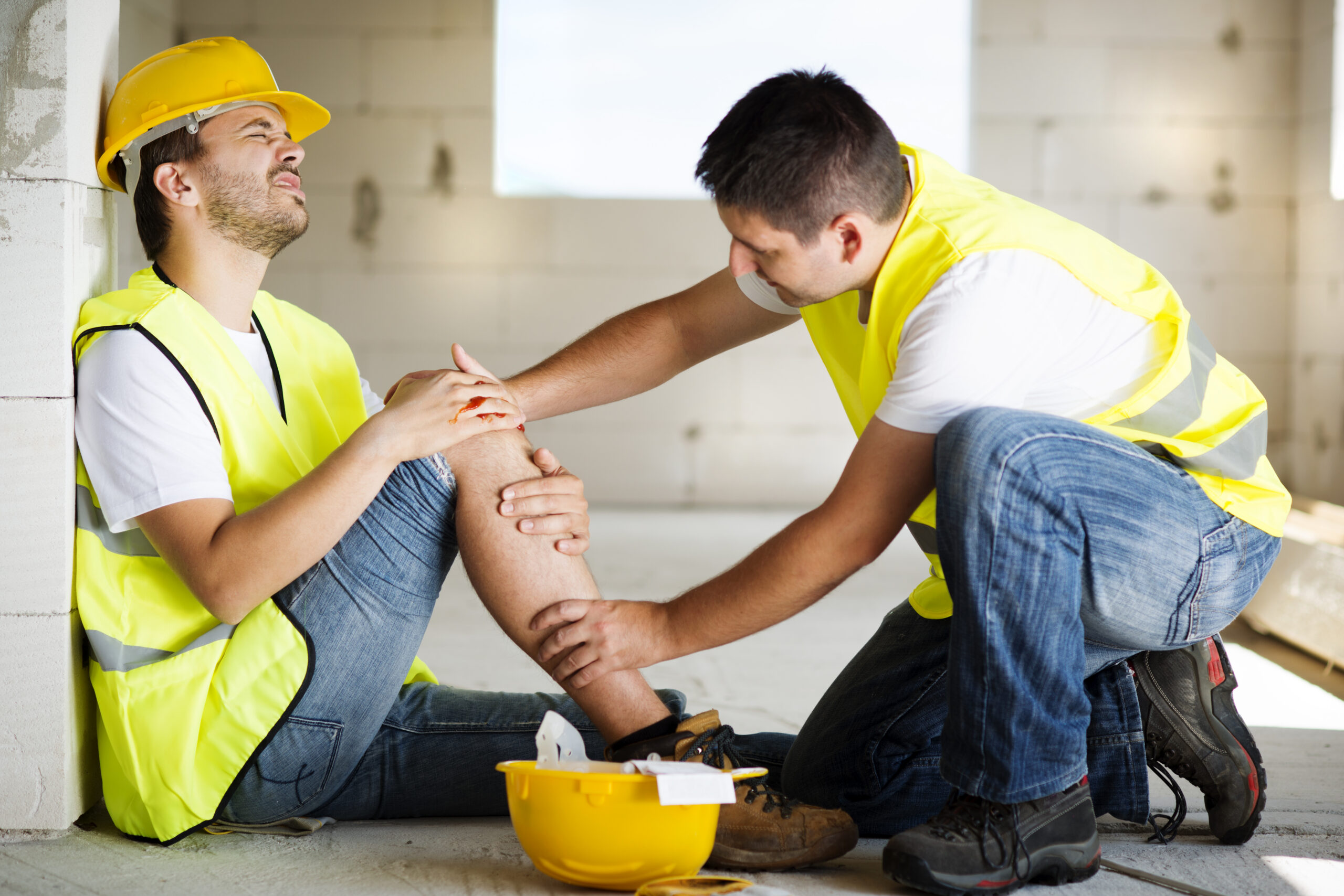 Construction worker in a yellow hard hat holding his injured knee while a colleague provides workplace injury care.